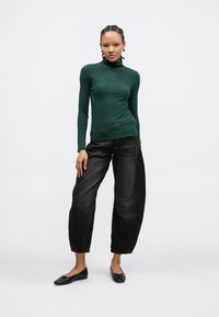 Πράσινο μακρυμάνικο τοπ με ψηλό γιακά, συνδυασμένο με ψηλόμεσα μαύρα τζιν παντελόνια. Το σύνολο έχει εφαρμοστή γραμμή και απαλή υφή.