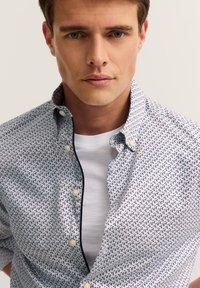 Junger Mann in einem weißen, gemusterten Hemd über einem schlichten weißen T-Shirt, mit neutralem Gesichtsausdruck und kurz geschnittenen braunen Haaren.