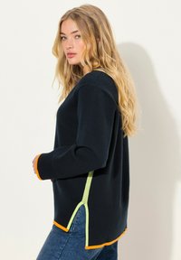 Donkerblauwe trui met oranje en groene accenten, een relaxte pasvorm, zijsplitten en geribbelde boorden, gecombineerd met blauwe jeans.