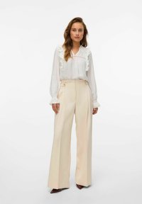 Witte blouse met ruches en lichtbeige hoge taillebroek. De outfit heeft een losse pasvorm en heeft knoopdetails.