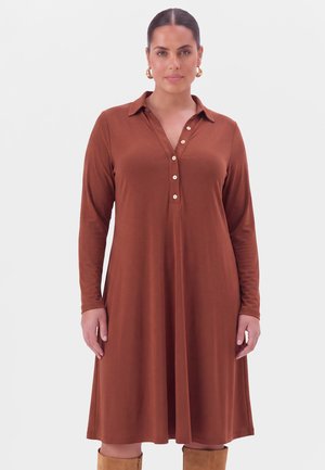 Femme portant une robe marron à manches longues boutonnée avec un col, des boucles d'oreilles anneaux dorés et des bottes beige hautes jusqu'aux genoux, debout sur un fond uni.