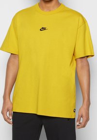 Žluté bavlněné tričko s kulatým výstřihom a krátkými rukávy. Obsahuje malé černé logo Nike na hrudi. Volný střih a hladká textura.