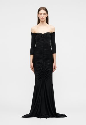 Donna che indossa un lungo abito nero scivolato sulle spalle con dettagli arricciati sulla parte frontale e maniche a tre quarti, in piedi davanti a uno sfondo semplice.