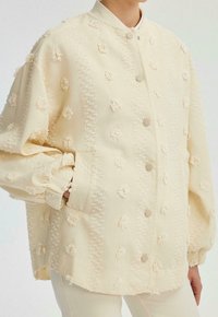 Veste beige clair avec broderie florale texturée, col montant et boutons-pression. Comprend des manches bouffantes et des poches latérales.
