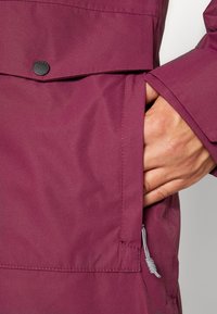 Main d'une personne dans la poche latérale d'une veste bordeaux avec rabat et bouton-pression noir, tirette de fermeture éclair rayée visible.