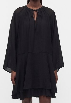 Femme portant une robe noire ample avec de longues manches larges et un col noué, ourlet en couches tombant au-dessus des genoux.