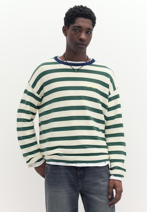 Ung mand med mørkt krøllet hår iført en grøn og hvid stribet langærmet skjorte, mørke jeans og en perlekrans, stående mod en ensfarvet baggrund.