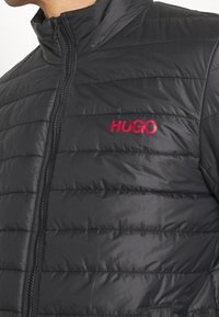 Svart pufferjacka med vertikalt sydd design, hög krage och fullängdsdragkedja. Har en röd "HUGO"-logotyp på bröstet.