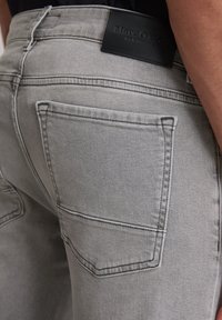 Gros plan sur une personne portant un jean gris clair avec une poche arrière visible et une étiquette de marque noire Marc O'Polo sur la ceinture.