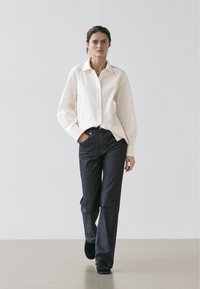 Camicia bianca con bottoni e maniche lunghe, abbinata a jeans a gamba larga di colore blu scuro. Texture complessivamente liscia e vestibilità rilassata; elementi di design semplici.