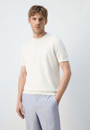 Jeune homme aux cheveux châtain clair, portant une chemise blanche à manches courtes et un pantalon bleu clair, debout avec une main dans la poche devant un fond uni.