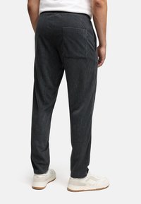 Mies, kajan tummanharmaat corduroy-housut, joissa on takatasku, ja valkoiset lenkkarit, seisoo yksinkertaista valkoista taustaa vasten.