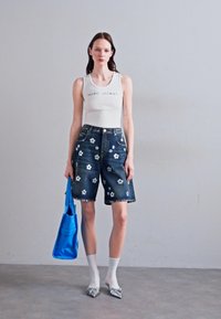 Shorts di denim con applicazioni floreali, abbinati a una maglietta senza maniche bianca. Tiene una borsa blu, indossa calzini bianchi e scarpe a punta argentate.