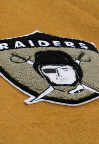 Black and beige embroidered Raiders logo on a golden fabric background, featuring a stylized pirate's face and crossed swords.