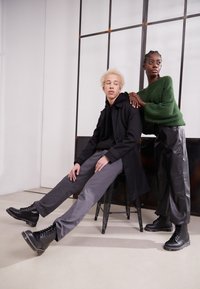 Botas de combate negras con cordones y suelas gruesas. Modelo con pantalones grises y abrigo negro sentado en un taburete, mientras otro lleva un suéter verde texturizado.