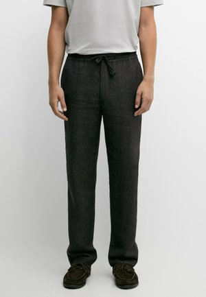 Uomo che indossa pantaloni scuri a gamba dritta con vita regolabile, abbinati a scarpe scamosciate scure e una camicia leggera a maniche corte.