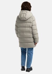 Lichte grijze pufferjas met capuchon, voorzien van een gewatteerd ontwerp en een verlengde snit. Gepaard met blauwe jeans en zwarte schoenen.