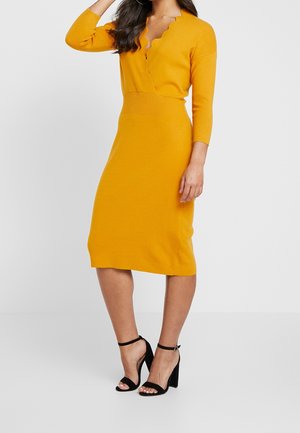 Robe en tricot jaune avec un décolleté en forme de vagues et des manches 3/4. Corsage ajusté et taille côtelée, longueur genou, associée à des sandales noires à talons hauts.