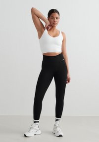 Ung kvinde iført en hvid crop top, sorte leggings med høj talje og hvide sneakers poserer med den ene arm bøjet bag hovedet.
