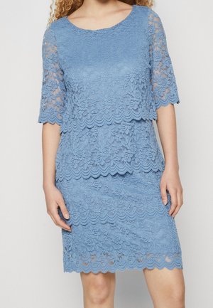 Femme portant une robe en dentelle bleu ciel à couches avec des manches trois-quarts et des bords festonnés, debout devant un fond clair uni.