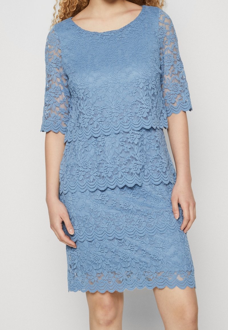 Femme portant une robe en dentelle bleu ciel à couches avec des manches trois-quarts et des bords festonnés, debout devant un fond clair uni.