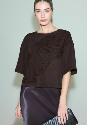 Nő barna, texturált, rövid ujjú felsőt és fényes sötét szoknyát visel, fekete szövött borítéktáskát tart egy egyszínű háttér előtt.