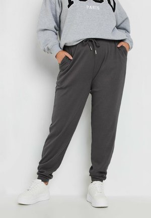 Graue, gerippte Jogginghosen mit elastischem Bund und Seitentaschen, kombiniert mit einem grauen Sweatshirt mit schwarzem Aufdruck. Weiße Sneaker.