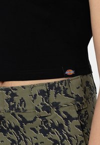Top court en coton noir associé à un pantalon en camouflage vert olive avec une surface texturée et un patch de marque sur la ceinture.
