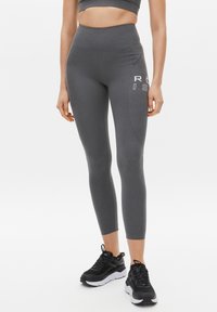 Graue, hochtaillierte Leggings aus elastischem Stoff mit einem seitlichen Logo in Weiß. Kombiniert mit schwarzen Sportschuhen mit strukturiertem Sohlenmaterial.
