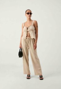 Beež crop top, millel on nöörkinnitus, koos lahtiste sobivate pükstega. Mudel kannab musti päikeseprille ja õlakotiga, seistes ühtlasel taustal.