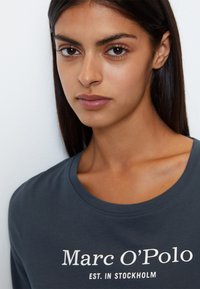 Dunkelgraues Baumwoll-T-Shirt mit rundem Halsausschnitt, auf dem "Marc O'Polo EST. IN STOCKHOLM" in Weiß aufgedruckt ist. Geschmeidige Textur und lässige Passform.