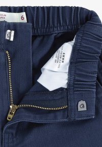 Pantalon à taille élastique bleu marine avec fermeture éclair ouverte, fermeture métallique à crochet et barre, et étiquette blanche de taille visible indiquant la taille 6 pour enfants de 5 à 6 ans.