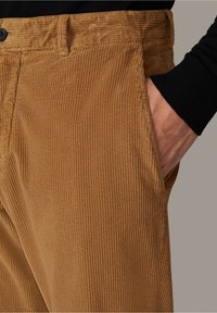 Close-up van een hand in de voorzak van een bruine corduroy broek, gedragen met een zwart geribbelde lange mouw shirt.