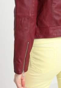 Giacca in pelle bordeaux che mette in evidenza il materiale texturizzato, design aderente, chiusura con zip laterale sulla manica, abbinata a pantaloni giallo pallido.