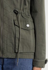 Olivgrön jacka med zip-öppning, diamant-quiltad yta, framficka med tryckknapp och dragsko i midjan. Ärmarna uppvikta.