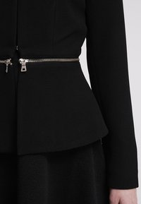 Schwarze strukturierte Jacke mit taillierter Taille, die ein silbernes Reißverschlussdetail und einen Peplum-Schnitt aufweist. Nahaufnahme von Stoff und Beschlägen.