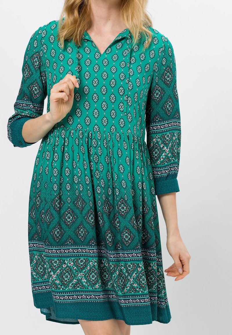 Femme portant une robe bleu sarcelle longueur genou avec des motifs géométriques et floraux mélangés, manches trois-quarts et un léger col en V.