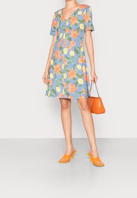 Vestido floral de mangas cortas, con tela azul, flores naranjas, limones amarillos y un escote en forma de V. La modelo sostiene un bolso naranja texturizado.