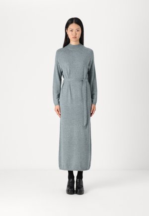 Long, light grey knitted dress with a high neck, long sleeves, and a tie waist. Flat, black platform shoes.