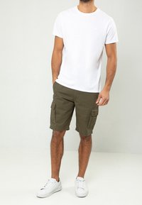 T-shirt blanc à col rond ; shorts cargo vert olive avec poches latérales ; baskets blanches. Tenue simple et décontractée sur un fond clair.
