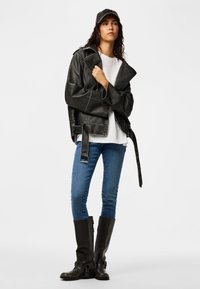 Femme portant une veste en cuir noire, une chemise blanche, un jean bleu, des bottes noires et une casquette noire, debout devant un fond uni.