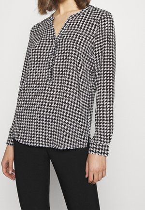 Vrouw draagt een zwart-witte blouse met pied-de-poule patroon, lange mouwen en een knopenhals, gecombineerd met een zwarte broek.