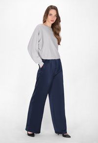 Lichtgrijze langesleeve top met een gathered taille, gecombineerd met marineblauwe wijde broekspijpen. Zwarte hoge hakken completeren de outfit.