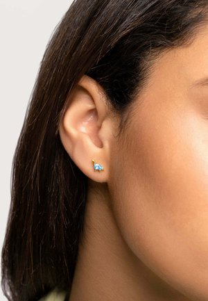 Goldfarbener, dinosaurierförmiger Ohrring mit einem blauen Edelstein-Akzent, am Ohr präsentiert. Nahaufnahme zeigt die Details des Designs und des Ohrs.