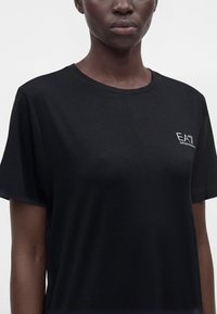 Άτομο που φοράει απλό μαύρο T-shirt με στρογγυλή λαιμόκοψη και μικρό λευκό λογότυπο EA7 Emporio Armani στο αριστερό στήθος, σε λευκό φόντο.
