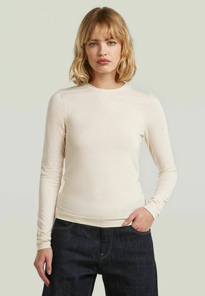 Vrouw met blond haar, gekleed in een beige shirt met lange mouwen dat in een donkerblauwe jeans is gestopt, met één hand in de zak, tegen een neutrale achtergrond.