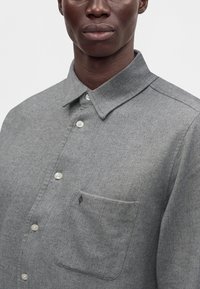 Homme portant une chemise grise à motif chevrons, avec poche poitrine et col, visible du cou jusqu'au milieu du torse.