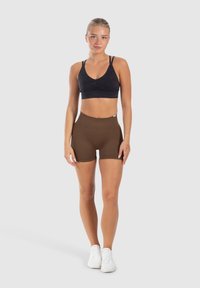 Zwarte sportbh met een V-hals en geplooide details, gecombineerd met bruine high-waisted shorts. Model draagt witte sportschoenen.