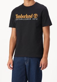 Černé bavlněné tričko s nápisem "Timberland" a rokem založení 1973 v oranžové barvě, se symbolem; klasický kulatý výstřih, krátké rukávy, běžný střih.