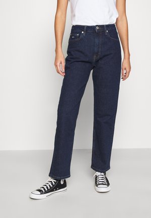 Persoon die donkerblauwe jeans met rechte pijpen, een wit T-shirt en zwarte hoge sportschoenen draagt, staand tegen een effen lichte achtergrond.